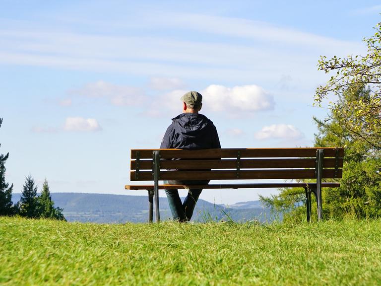 Ein älterer Herr sitzt mit dem Rücken zum Betrachter auf einer Parkbank mit Blick in die Ferne.