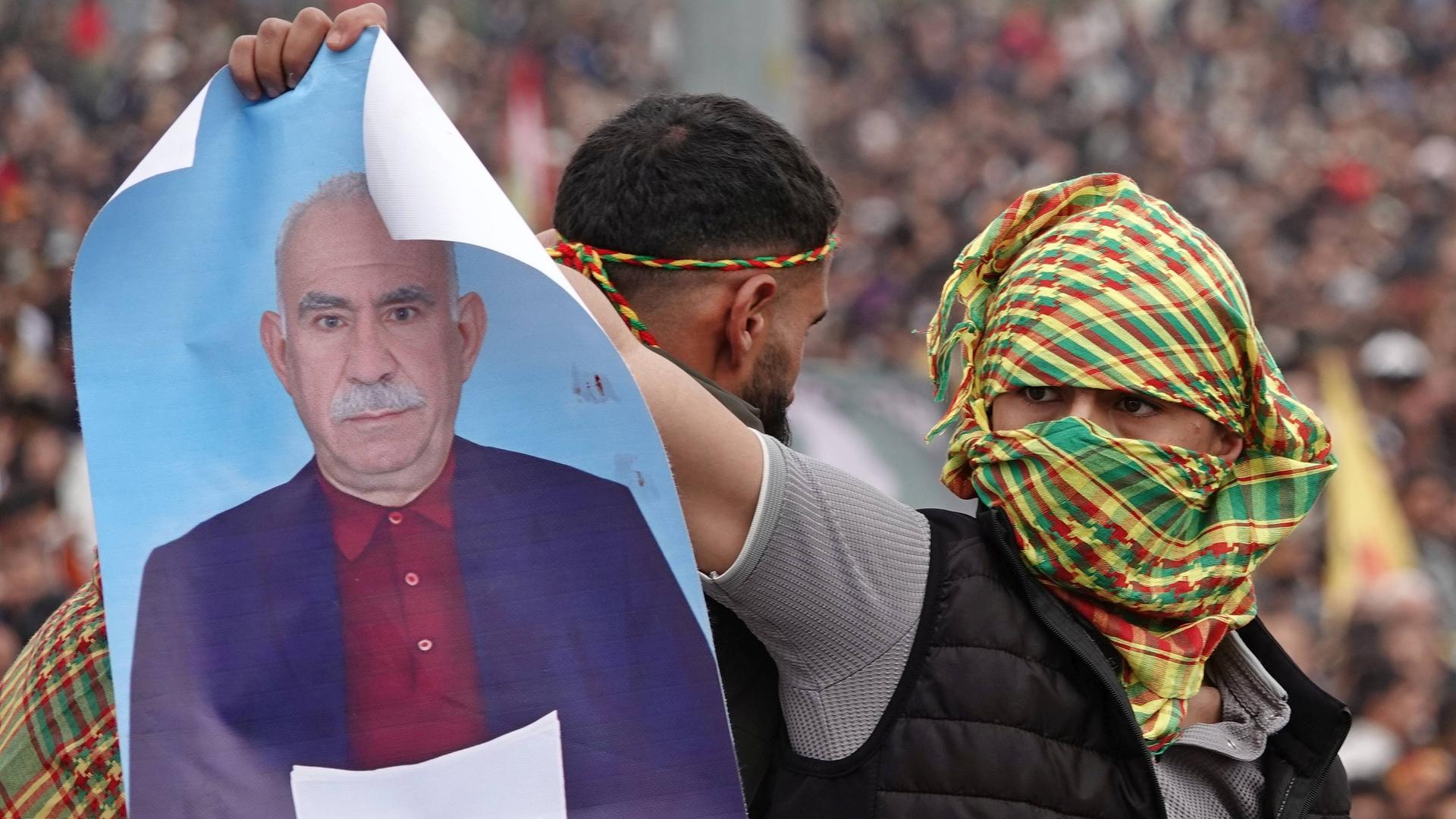 Zu sehen ist ein mit buntem Tuch vermummter Demonstrant. Dieser hält ein Poster hoch, das Abdullah Öcalan zeigt. 