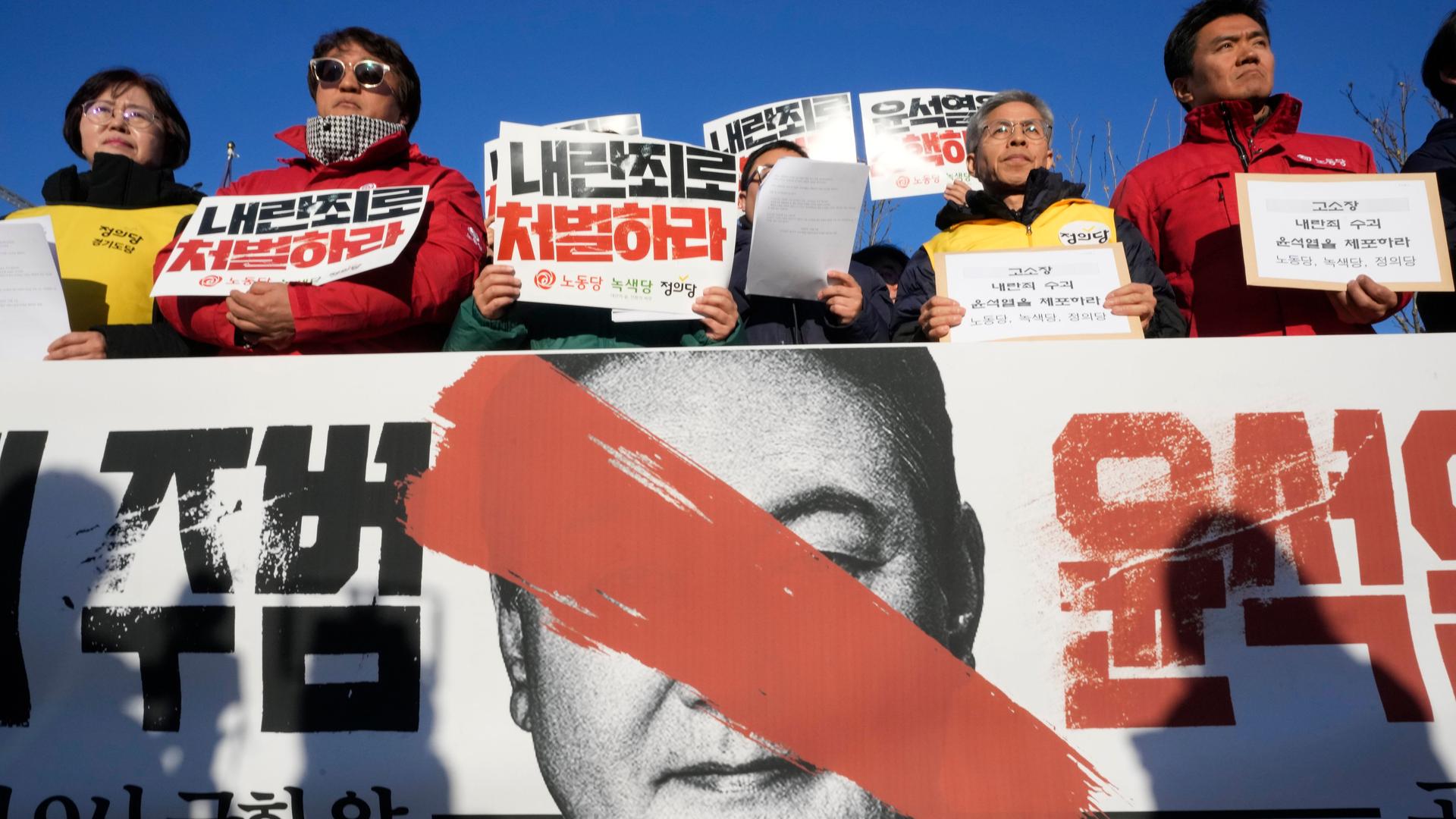 Das Bild zeigt Demonstranten, die mit Schildern und Plakaten den Rücktritt des südkoreanischen Präsidenten Yoon Suk Yeol fordern. 