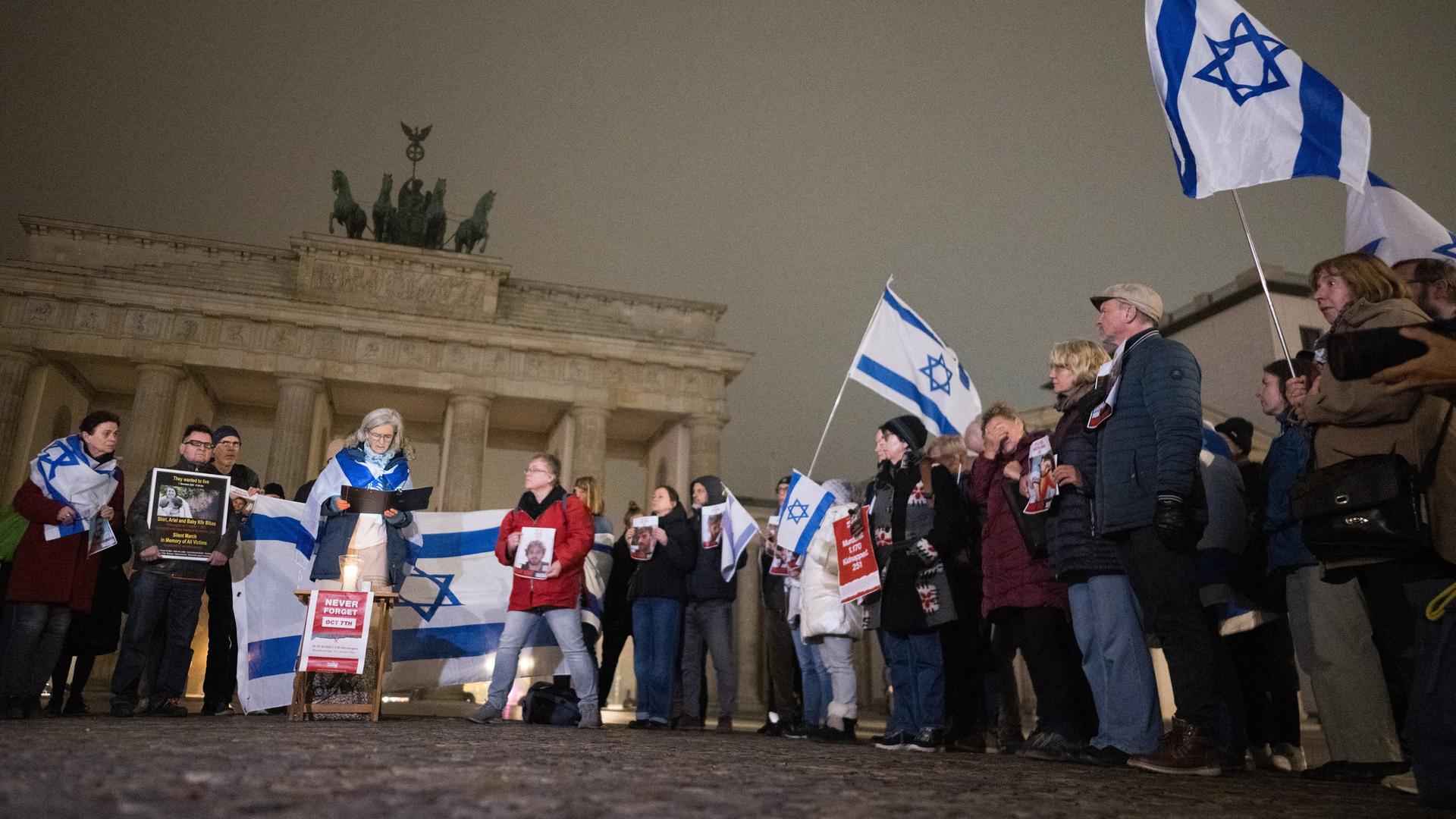 Menschen stehen im Morgengrauen am Brandenburger Tor und lesen die Namen der Opfer vom 7. Oktober 2023 vor. Einige von ihnen tragen Israelfahnen