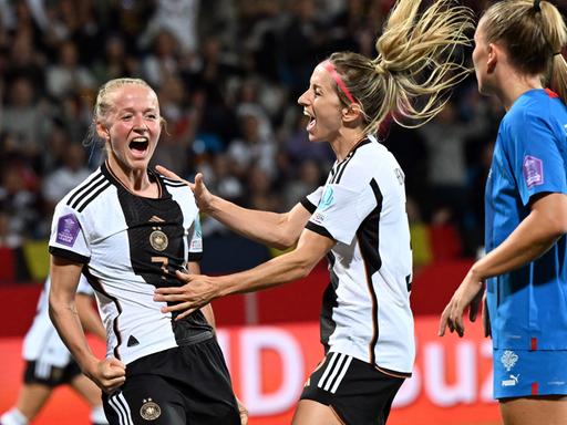 Zwei Fußballerinnen im Deutschland-Trikot jubeln; eine isländische Gegenspielerin beobachtet sie dabei.