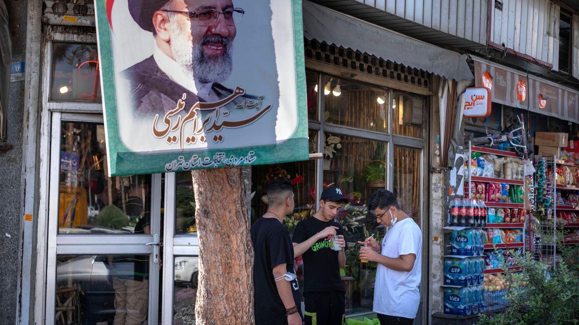 Drei Jugendliche stehen vor einem Lebensmittelgeschäft neben einem Baum mit dem Plakat des iranischen Präsidenten Raisi.