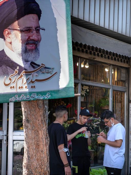 Drei Jugendliche stehen vor einem Lebensmittelgeschäft neben einem Baum mit dem Plakat des iranischen Präsidenten Raisi.
