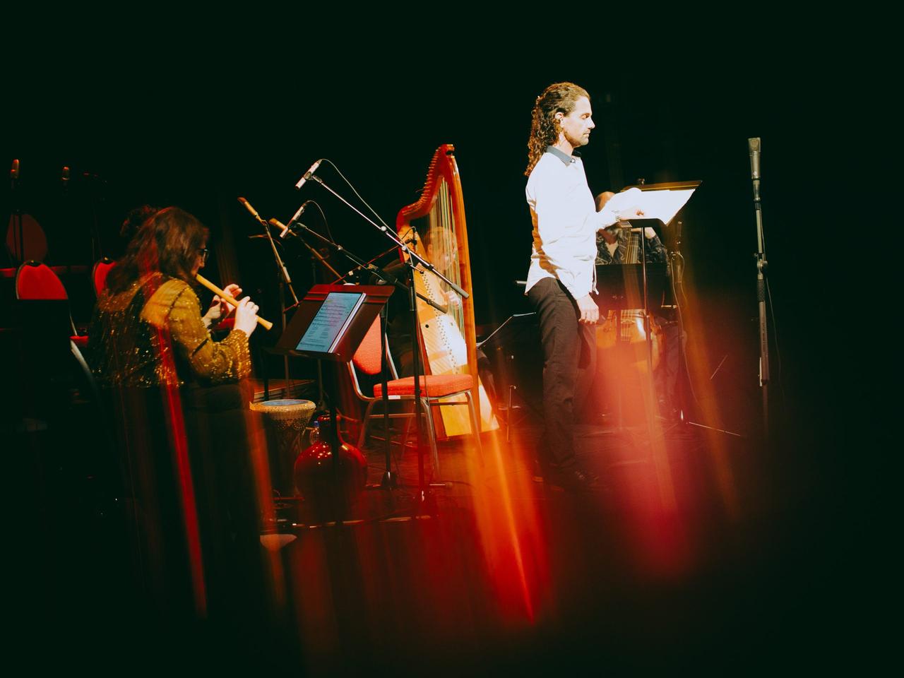 Ein Sänger steht vor einem Mikrofon, während hinter ihm eine Harfe und eine Flötenspielerin erkennbar ist.