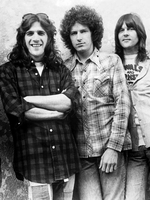 Auf dem Schwarzweiß-Bild stehen die Bandmitglieder der Eagles, Bernie Leadon, Glenn Frey, Don Henley, Randy Meisner und Don Felder (v.l.n.r.), lächelnd nebeneinander vor einer Wand.
