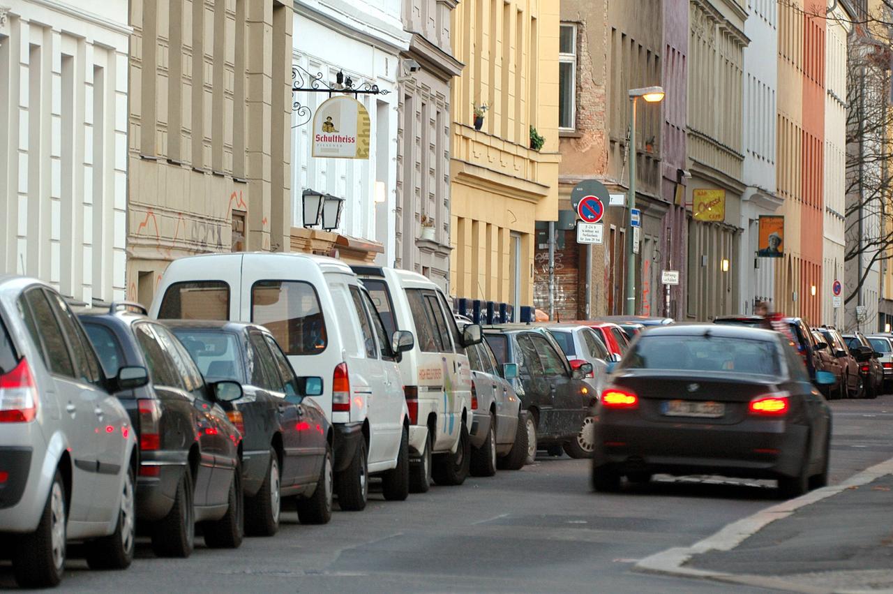 Viele Autos parken am Straßenrand vor Häusern. Auf der Straße fährt ein Auto.