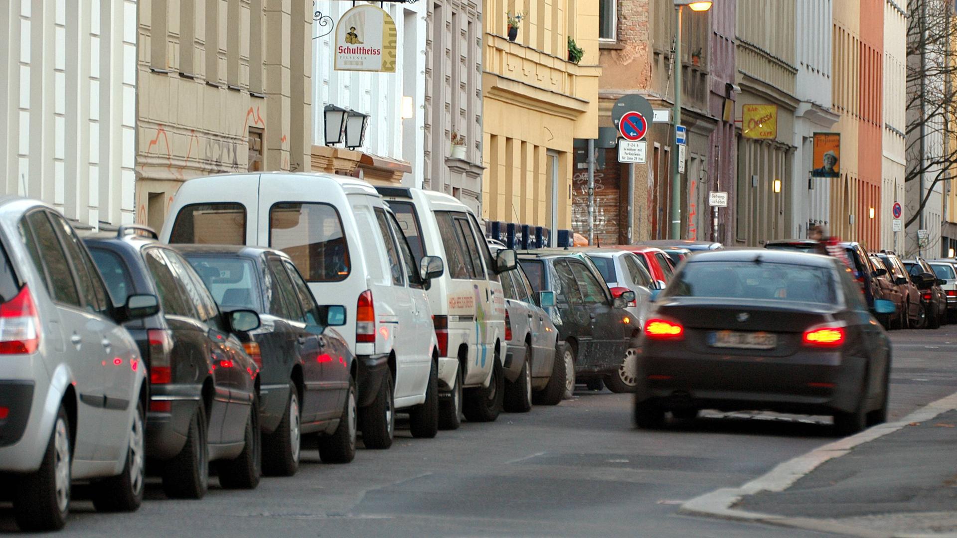 Viele Autos parken am Straßenrand vor Häusern. Auf der Straße fährt ein Auto.