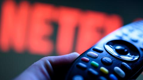 Eine Hand hält eine Fernbedienung und der Schriftzug Netflix im Hintergrund. 