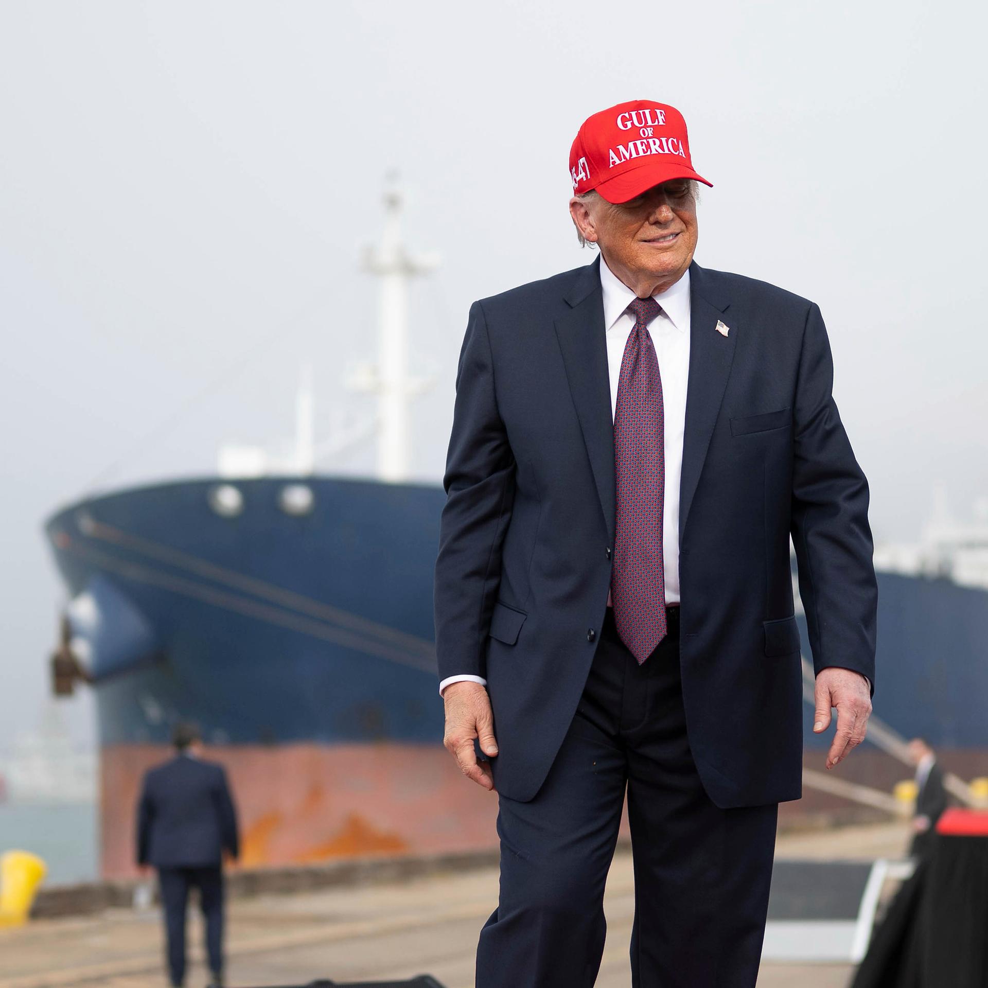US-Präsident Donald Trump steht an einem Hafen, hinter ihm ist ein großes Schiff zu sehen. Er trägt eine rotte Kappe mit der Aufschrift "Gulf of America"