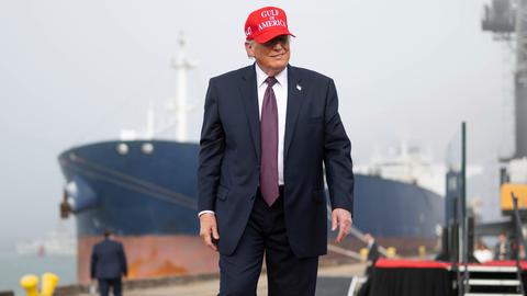 US-Präsident Donald Trump steht an einem Hafen, hinter ihm ist ein großes Schiff zu sehen. Er trägt eine rote Kappe mit der Aufschrift "Gulf of America".