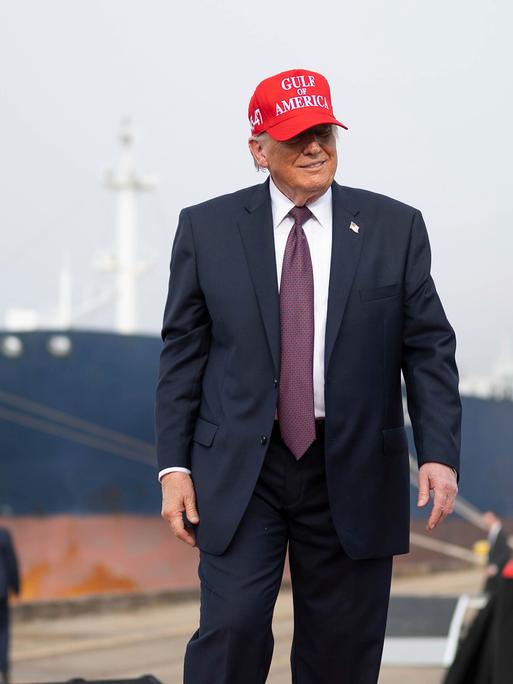 US-Präsident Donald Trump steht an einem Hafen, hinter ihm ist ein großes Schiff zu sehen. Er trägt eine rotte Kappe mit der Aufschrift "Gulf of America"