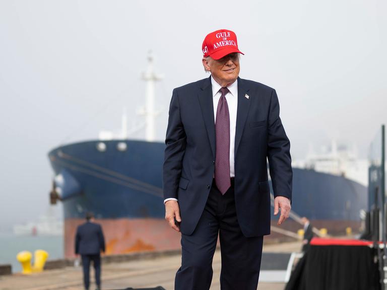 US-Präsident Donald Trump steht an einem Hafen, hinter ihm ist ein großes Schiff zu sehen. Er trägt eine rotte Kappe mit der Aufschrift "Gulf of America"