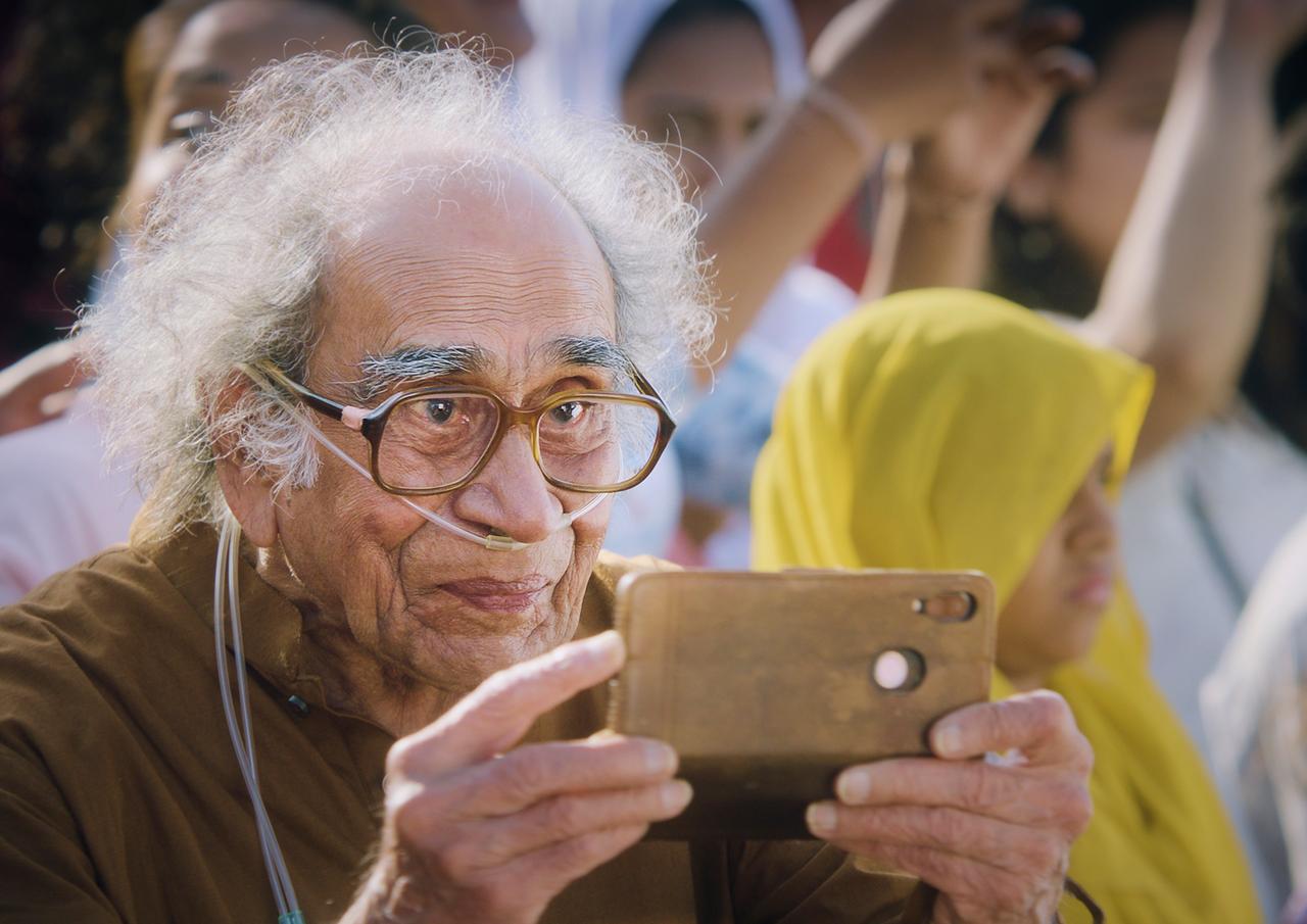 Ein alter Mann mit weißen, lockigen Haare, dem unterhalb der Nase einen Schlauch durchs Gesicht läuft, blickt auf ein Smartphone.
