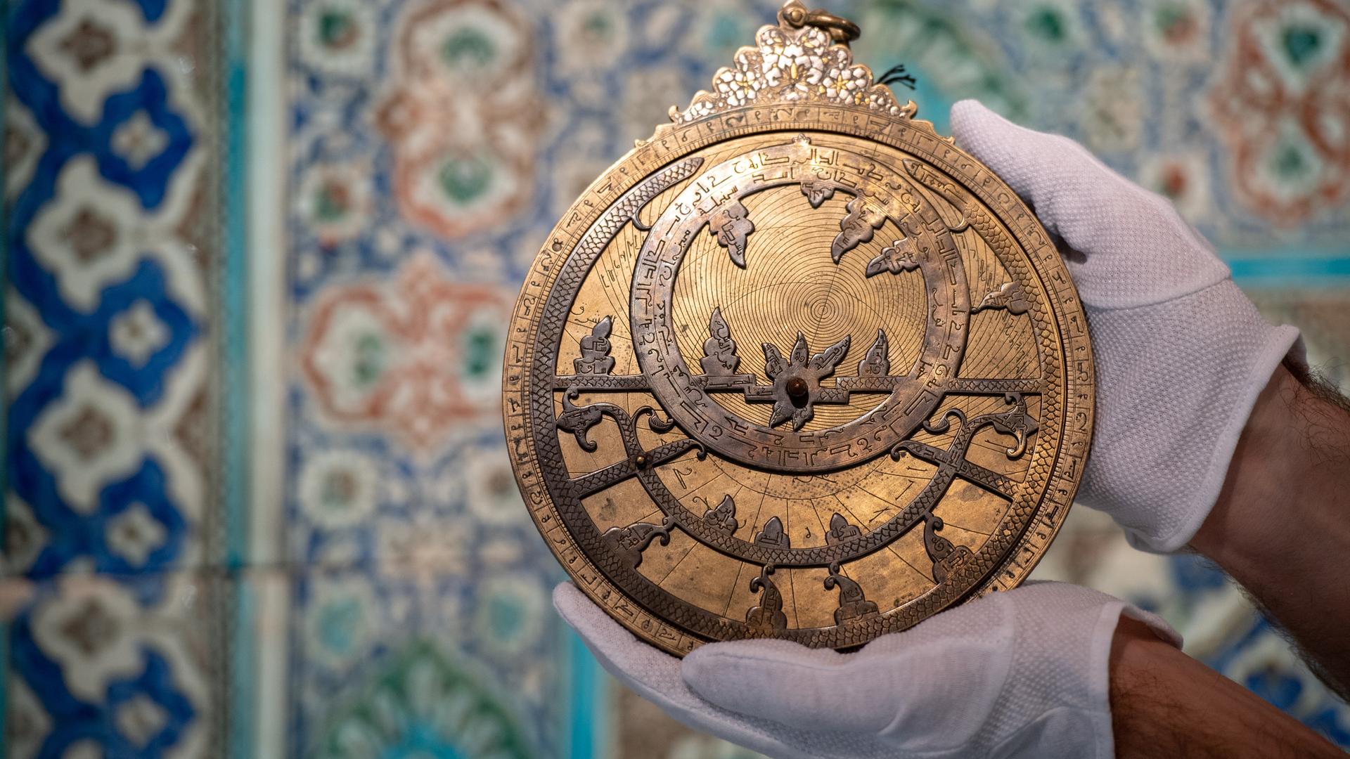 Weiß behandschuhte Hände halten ein Astrolabium in die Kamera, eine reich verzierte Scheibe aus vergoldetem Messing.