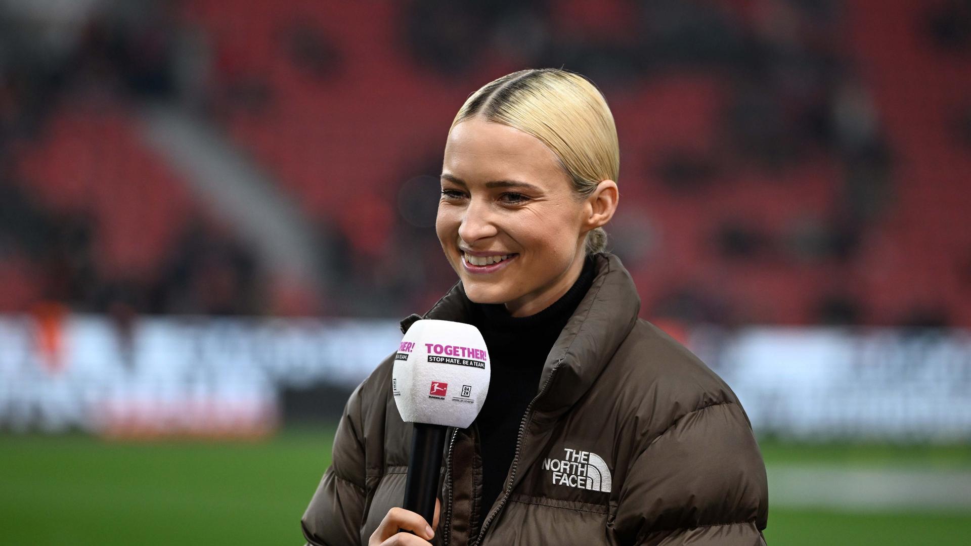 Die Sportmoderatorin Lena Cassel am Mikrofon bei einem Bundesligaspiel im Stadion. Sie trägt einen Rollkragenpullover und eine braune Daunenjacke. Die Sportmoderatorin Lena Cassel am Mikrofon bei einem Bundesligaspiel im Stadion. Sie trägt einen Rollkragenpullover und eine braune Daunenjacke.