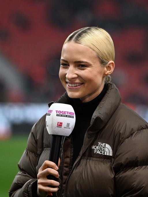 Die Sportmoderatorin Lena Cassel am Mikrofon bei einem Bundesligaspiel im Stadion. Sie trägt einen Rollkragenpullover und eine braune Daunenjacke.