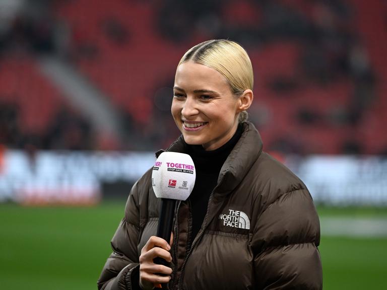 Die Sportmoderatorin Lena Cassel am Mikrofon bei einem Bundesligaspiel im Stadion. Sie trägt einen Rollkragenpullover und eine braune Daunenjacke.
