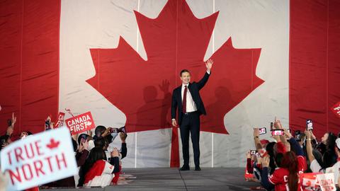 Pierre Poilievre steht bei einer Rally auf der Bühne und winkt. Hinter ihm an der Wand prangt ein riesiges stilisiertes rotes Ahornblatt.
