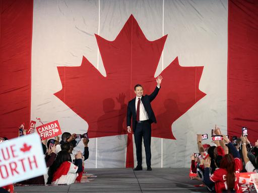Pierre Poilievre steht bei einer Rally auf der Bühne und winkt. Hinter ihm an der Wand prangt ein riesiges stilisiertes rotes Ahornblatt.