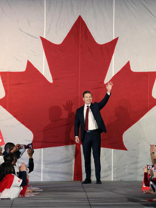 Pierre Poilievre steht bei einer Rally auf der Bühne und winkt. Hinter ihm an der Wand prangt ein riesiges stilisiertes rotes Ahornblatt.