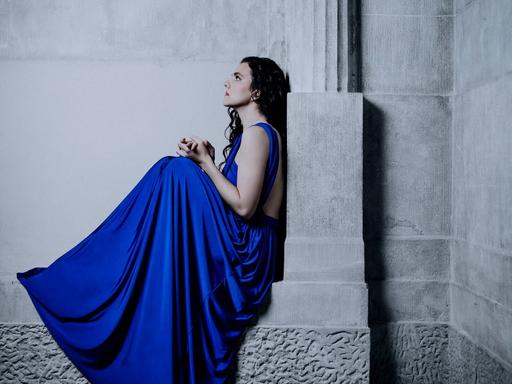 Die Pianistin sitzt in einem tiefblauem Kleid auf einem Gebäudevorsprung mit Säule, der an eine griechische Tempelanlage erinnert.