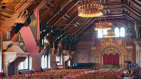 Blick in den mit Holz ausgetäfelten und mit vielen Fenstern bestückte lange Saal, in dem viele Zuschauerstühle stehen.