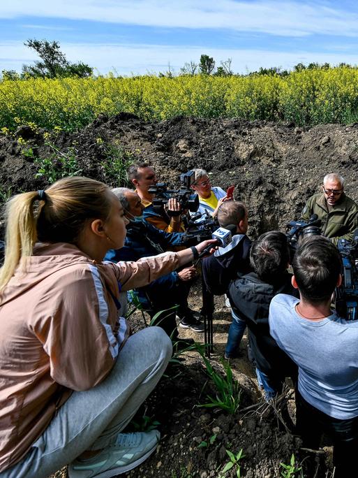 Medienvertreter sitzen im Kreis um einen ukrainischen Militär im Feld. 