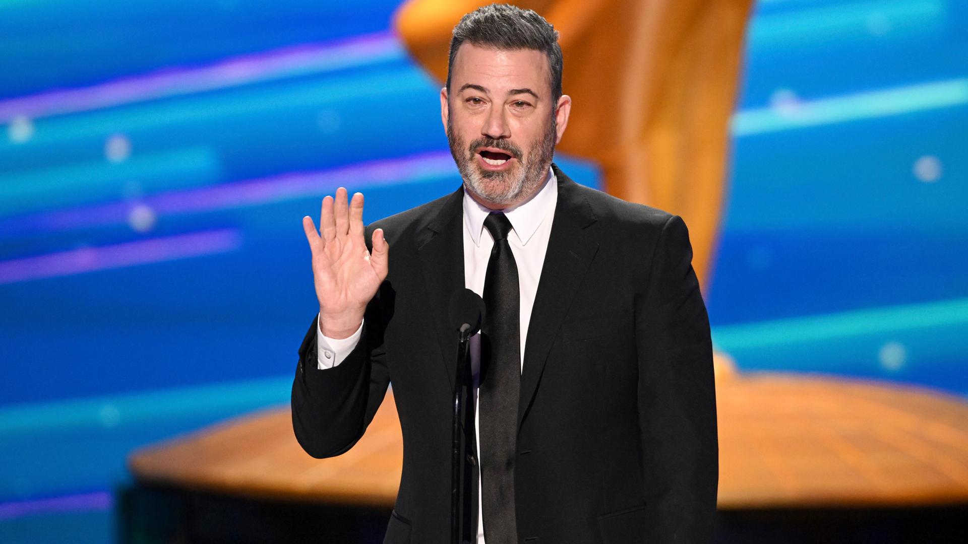 Jimmy Kimmel steht im schwarzen Anzug auf einer Bühne vor einem Mikrofon. Er hebt seine rechte Hand. Im Hintergrund ist viel Gold und Blau.