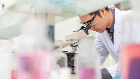 Eine Person im Schutzanzug und mit Schutzbrille steht in einem Labor und schaut durch ein Mikroskop. Eine Person im Schutzanzug und mit Schutzbrille steht in einem Labor und schaut durch ein Mikroskop.