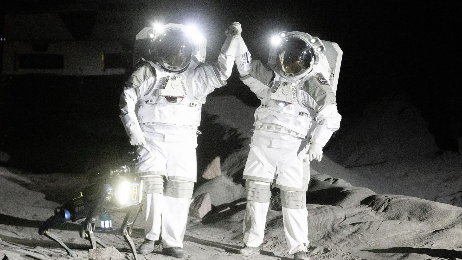 Die beiden Astronauten in weißen Raumanzügen stehen auf der modellierten Mondoberfläche und halten gemeinsam eine Hand hoch. Neben ihnen ein Mondroboter. Die beiden Astronauten in weißen Raumanzügen stehen auf der modellierten Mondoberfläche und halten gemeinsam eine Hand hoch. Neben ihnen ein Mondroboter.