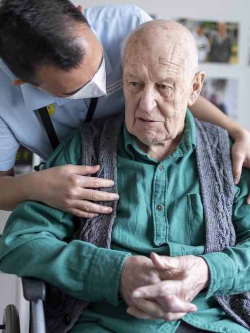 Ein Altenpfleger beugt sich über die Schulter eines alten, sitzenden Mannes und wendet sich ihm zu.