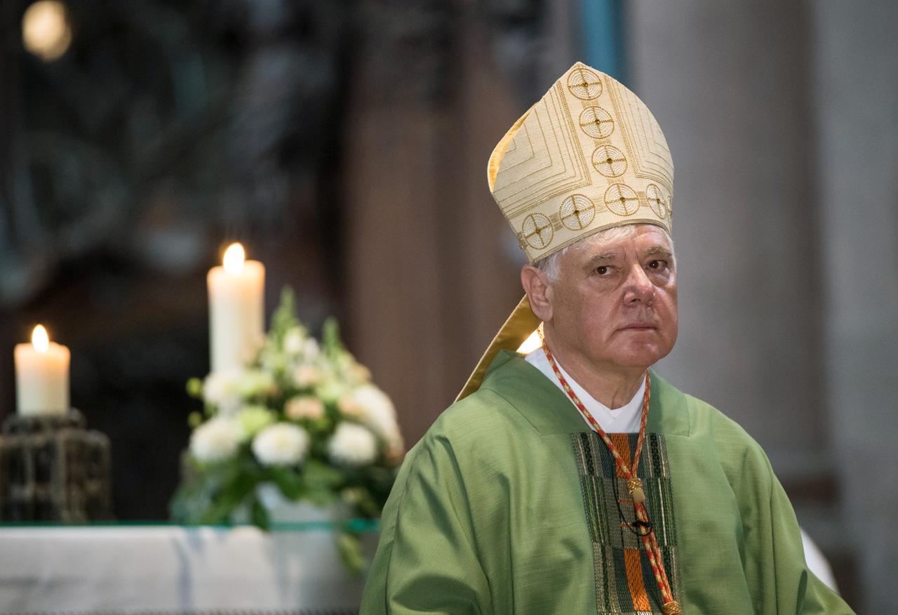 Kardinal Müller mit der Mitra auf dem Kopf guckt skeptisch in die Kamera. Im Hintergrund ist eine angezündete Kerze zu sehen.