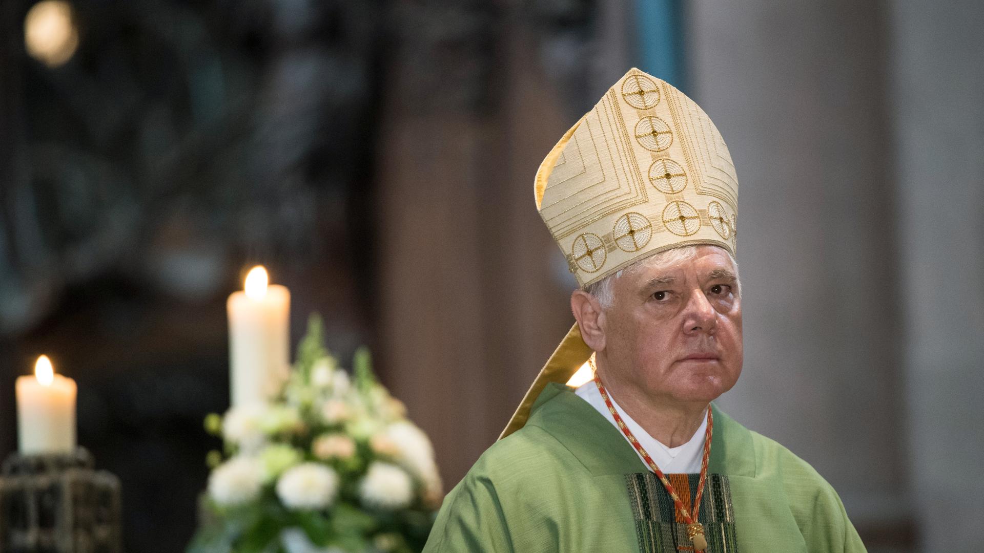Kardinal Müller mit der Mitra auf dem Kopf guckt skeptisch in die Kamera. Im Hintergrund ist eine angezündete Kerze zu sehen.