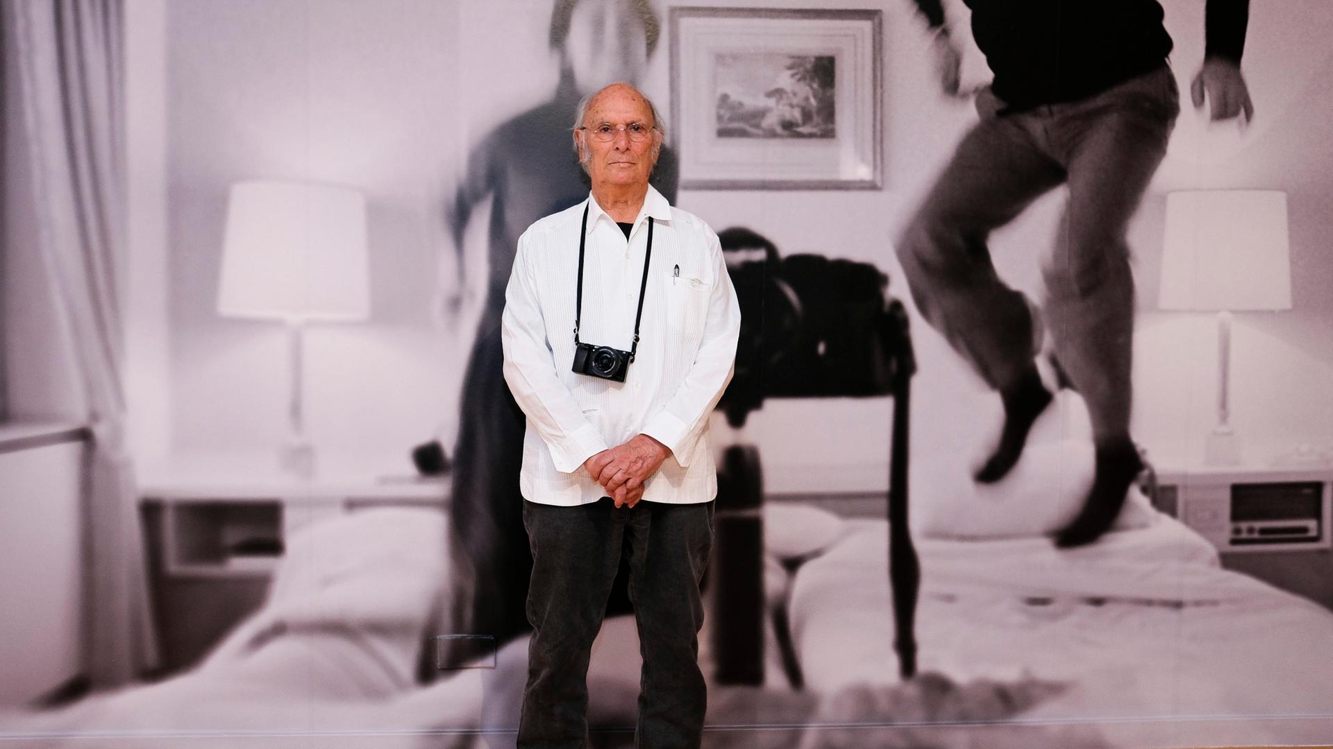 Carlos Saura steht vor einer riesigen Schwarz-Weiß-Fotografie, er trägt eine Kamera um den Hals.