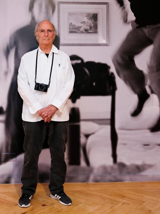 Carlos Saura steht vor einer riesigen Schwarz-Weiß-Fotografie, er trägt eine Kamera um den Hals.