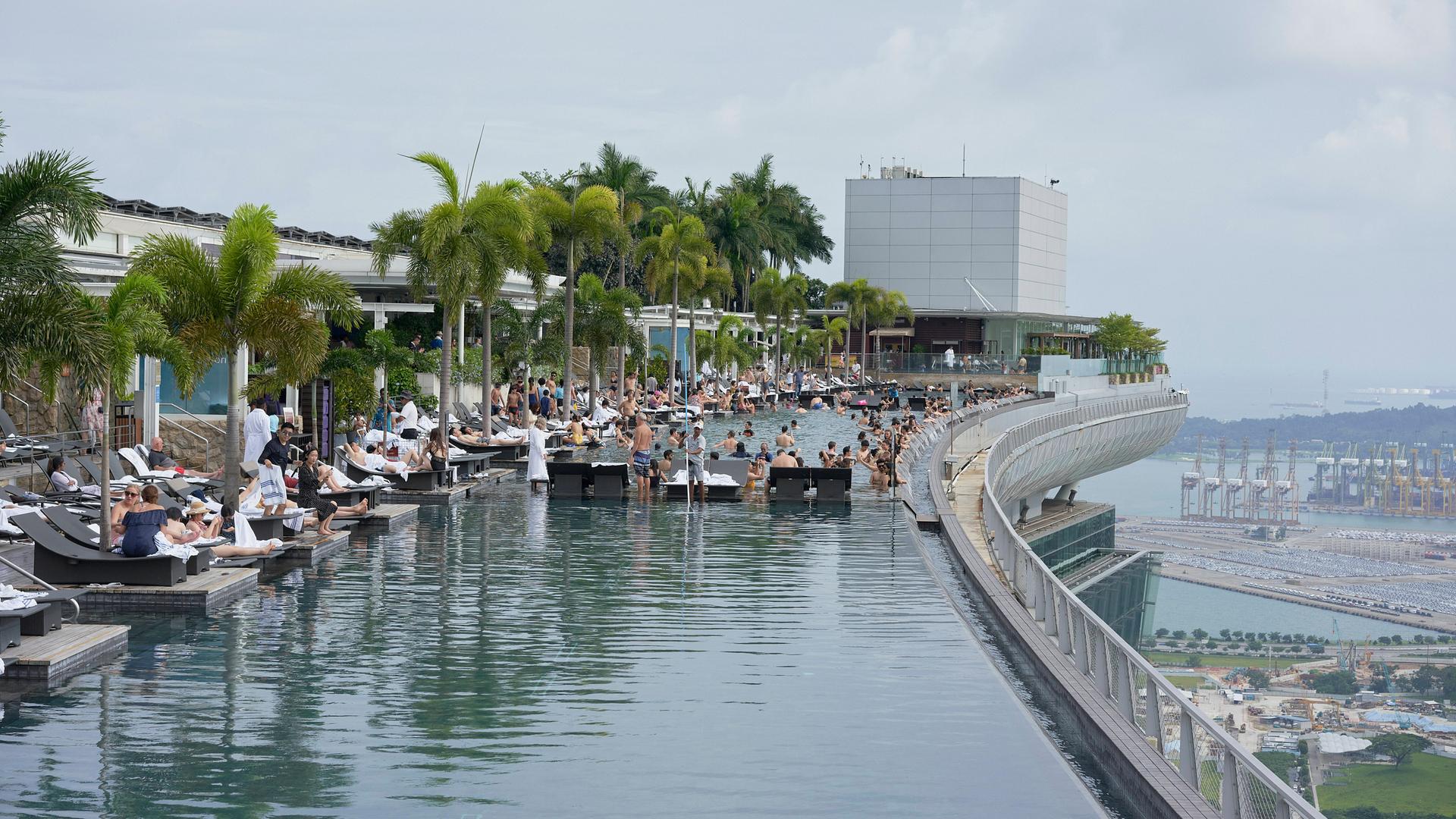 Blick auf die Dachterrasse eines Luxus-Hotels in Singapur. Menschen liegen an einem Pool, es sind diverse Palmen zu sehen.
Blick auf die Dachterrasse eines Luxus-Hotels in Singapur. Menschen liegen an einem Pool, es sind diverse Palmen zu sehen.