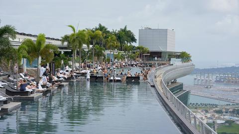 Blick auf die Dachterrasse eines Luxus-Hotels in Singapur. Menschen liegen an einem Pool, es sind diverse Palmen zu sehen.
Blick auf die Dachterrasse eines Luxus-Hotels in Singapur. Menschen liegen an einem Pool, es sind diverse Palmen zu sehen.