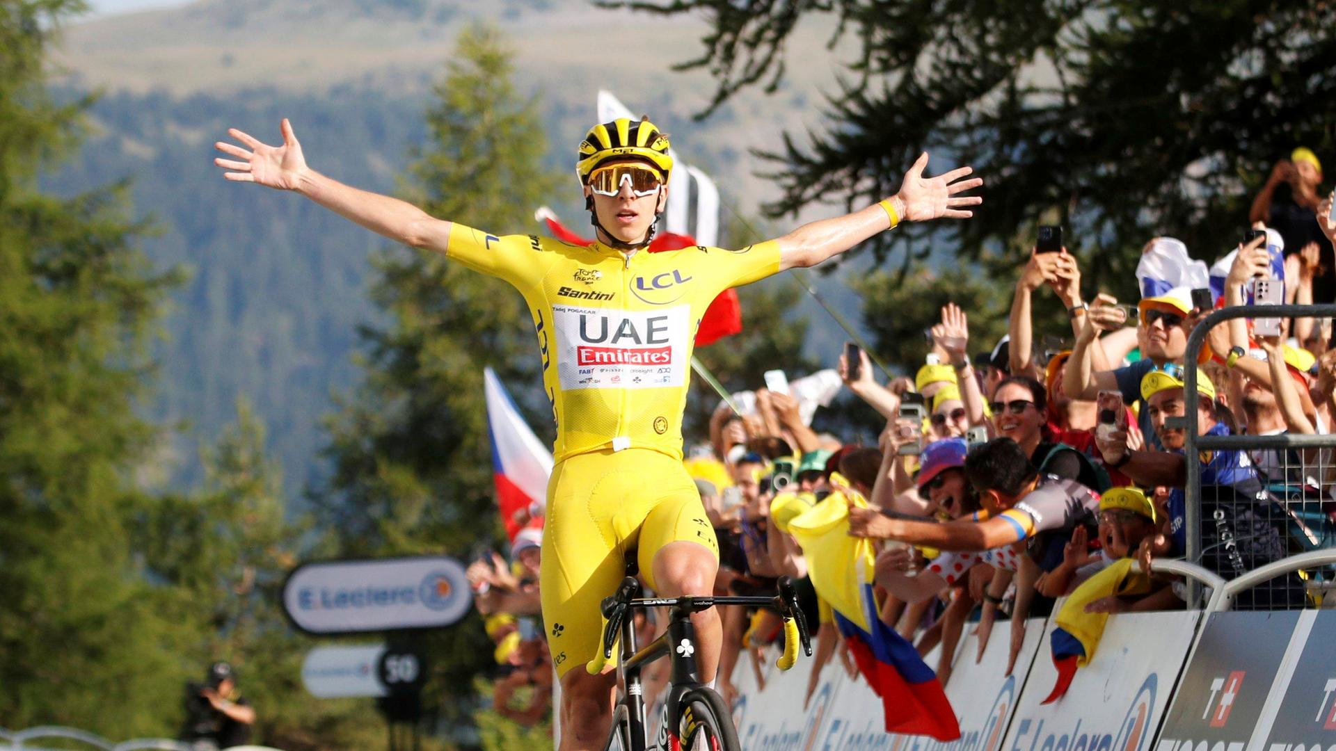 Radsprofi Tadej Pogacar fährt mit ausgestreckten Armen durch das Ziel der 20. Etappe der Tour de France 2024. Er trägt das gelbe Trikot des Geamtführenden. Hinter ihm jubeln ihm Fans zu.