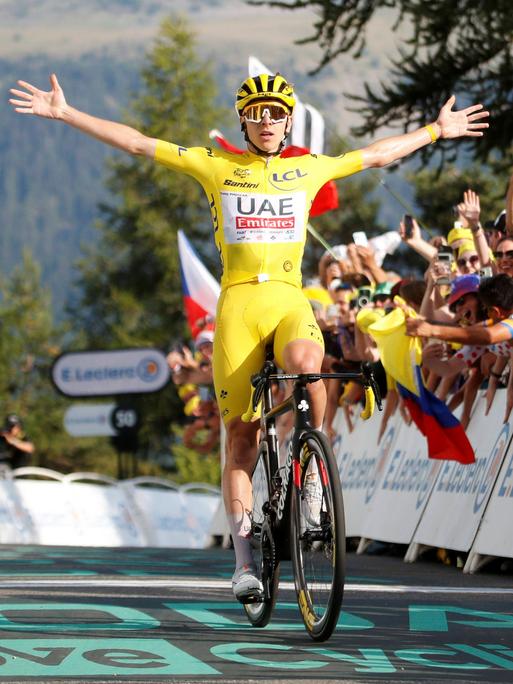 Radsprofi Tadej Pogacar fährt mit ausgestreckten Armen durch das Ziel der 20. Etappe der Tour de France 2024. Er trägt das gelbe Trikot des Geamtführenden. Hinter ihm jubeln ihm Fans zu.