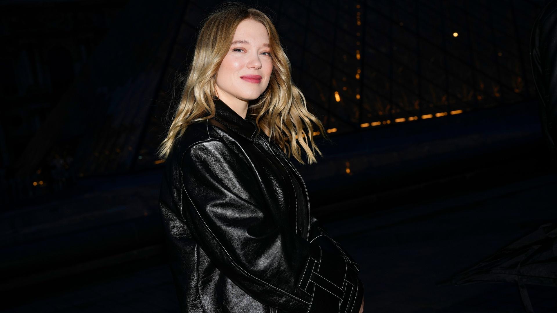 Die französische Schauspielerin Lea Seydoux steht in einem schwarzen Mantel vor schwarzem Hintergrund und lächelt in die Kamera.