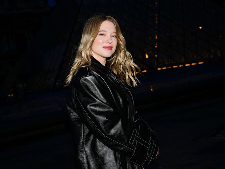 Die französische Schauspielerin Lea Seydoux steht in einem schwarzen Mantel vor schwarzem Hintergrund und lächelt in die Kamera. 