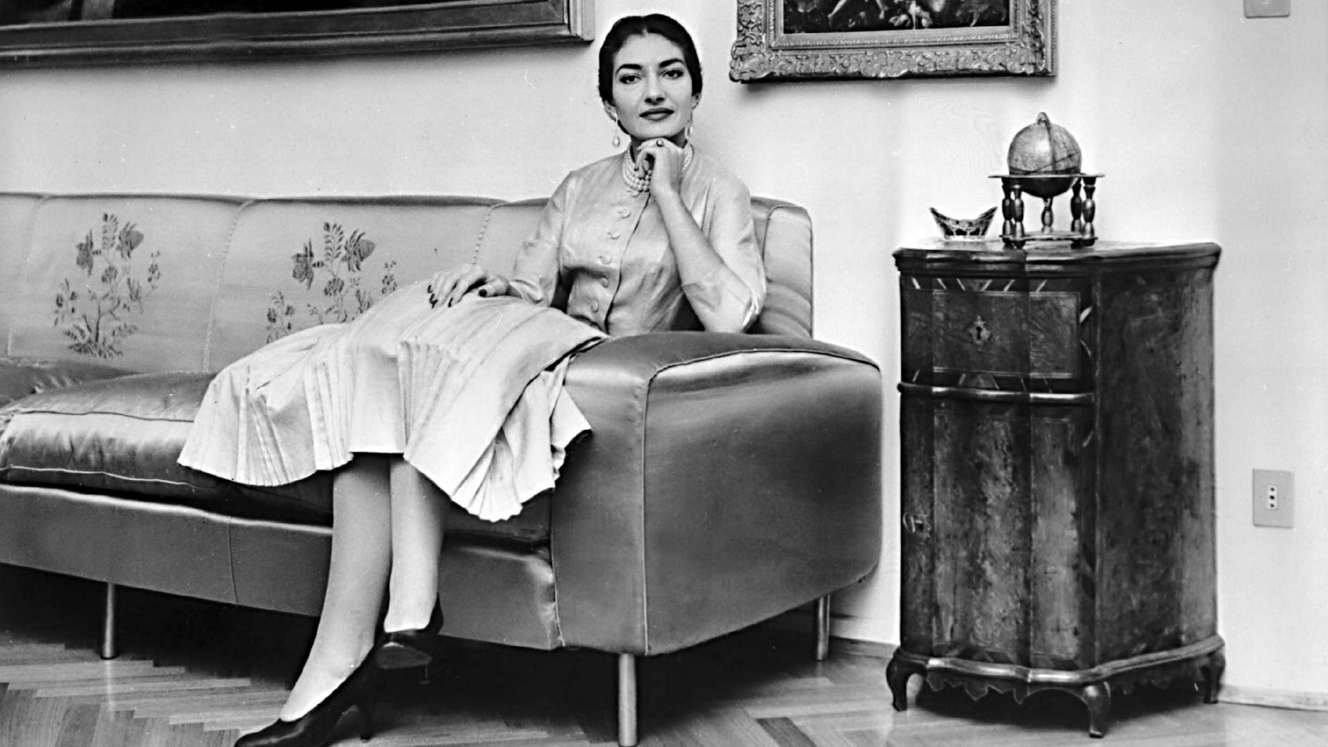 Maria Callas sitzt auf einem Sofa, über ihr hängen Gemälde. Sie trägt ein helles Kostüm und schaut in die Kamera.