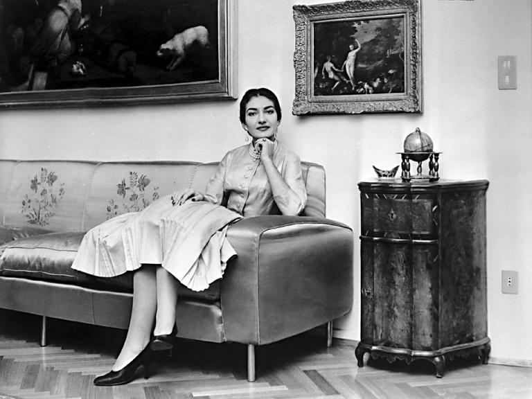 Maria Callas sitzt auf einem Sofa, über ihr hängen Gemälde. Sie trägt ein helles Kostüm und schaut in die Kamera.