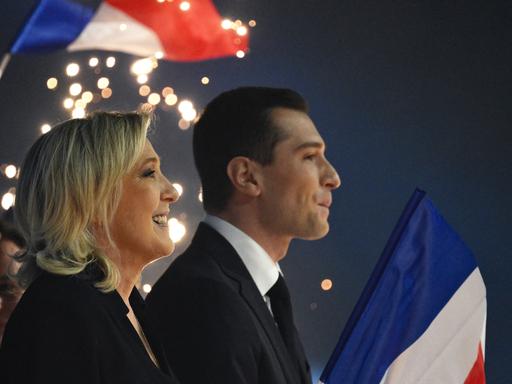 Marine Le Pen und Jordan Bardella stehen nebeneinander singend auf einer Bühne. Im Hintergrund sind französische Flaggen und Feuerwerk zu sehen.