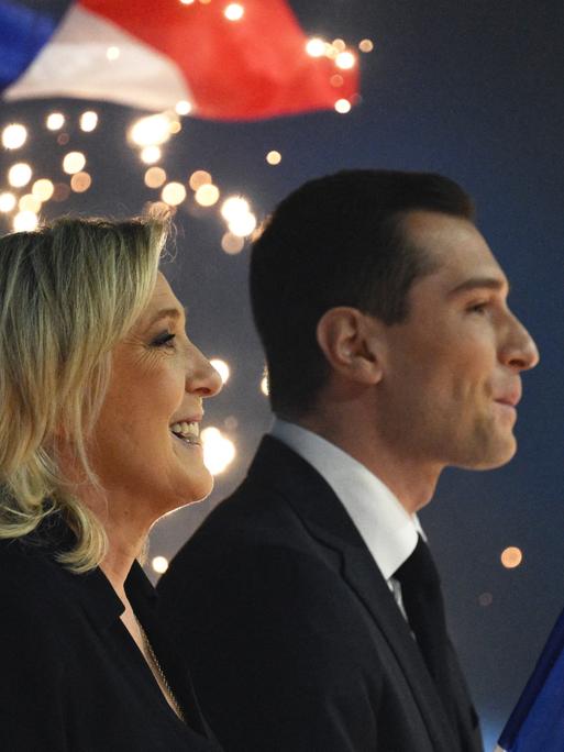 Marine Le Pen und Jordan Bardella stehen nebeneinander singend auf einer Bühne. Im Hintergrund sind französische Flaggen und Feuerwerk zu sehen.