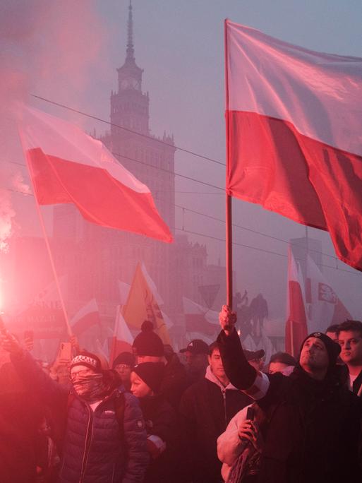 An einem nebligen Tag schwenken Protestierende polnische Flaggen. Im Hintergrund ist die Silhouette des Warschauer Kulturpalastes zu sehen.