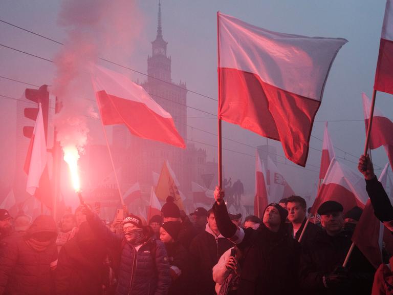 An einem nebligen Tag schwenken Protestierende polnische Flaggen. Im Hintergrund ist die Silhouette des Warschauer Kulturpalastes zu sehen.