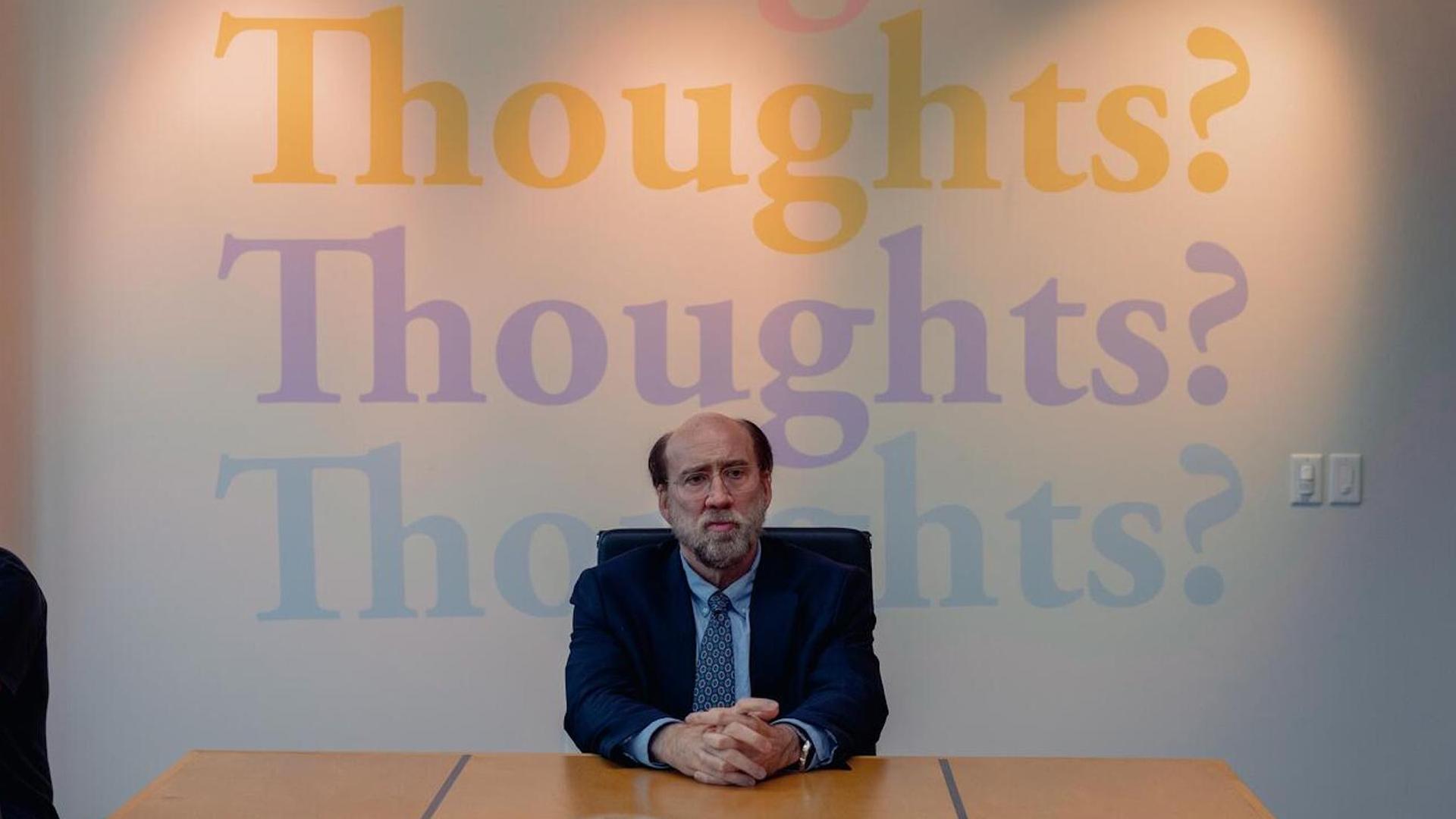 Im Still aus "Dream Scenario" sitzt Nicolas Cage mit nachdenklichem Gesichtsausdruck an einem Tisch. Hinter ihm steht in großen, bunten Buchstaben das Wort "Thoughts?" an der Wand.