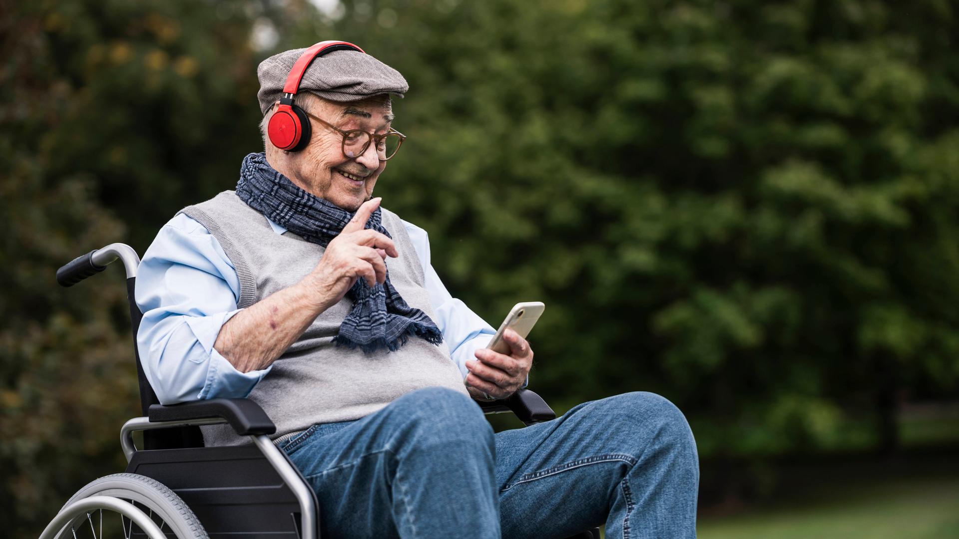 Ein Mann sitzt lächelnd im Rollstuhl und hat Kopfhörer auf. Im Hintergrund grüne Bäume und Büsche. Ein Mann sitzt lächelnd im Rollstuhl und hat Kopfhörer auf. Im Hintergrund grüne Bäume und Büsche.