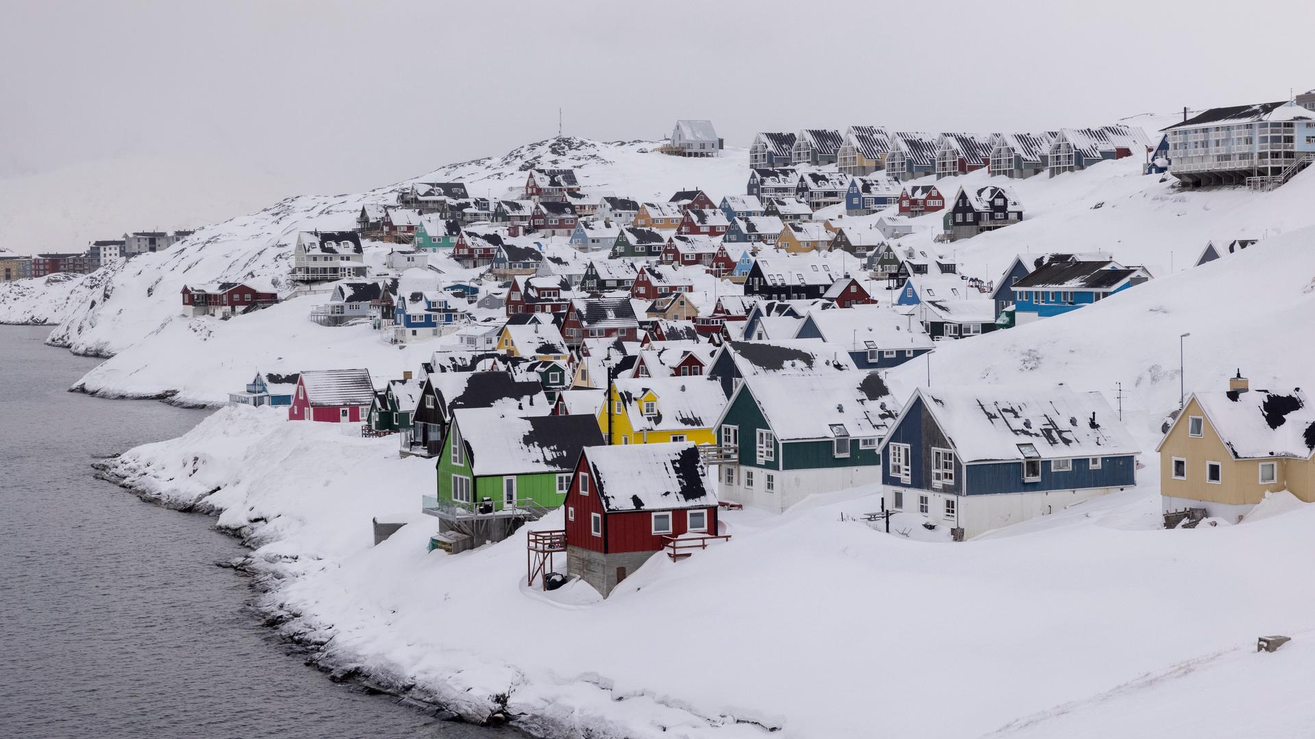 Bunte Holzhäuser stehen dicht gedrängt in der tief verschneiten Hauptstadt Nuuk. Links ist Gewässer zu sehen. 